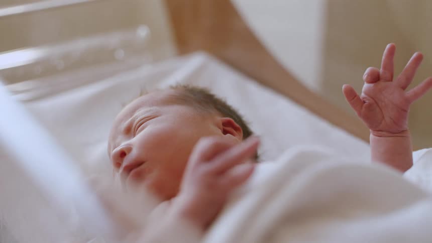 Newborn baby moving hands while resting in crib inside a nursery at hospital