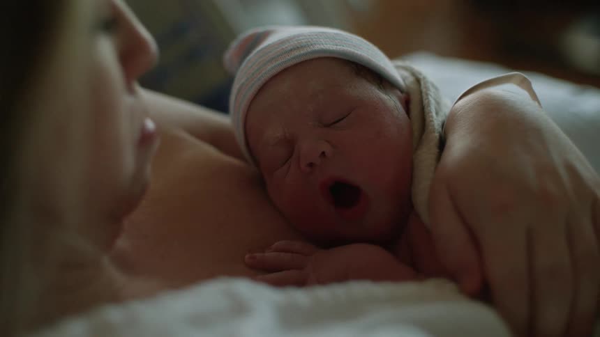 A new mom looks down at her yawning newborn held snuggly against her chest in hospital