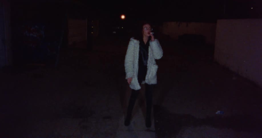 Woman with curly hair glasses black leather jacket and white feather jacket smokes a cigarette in the street at night