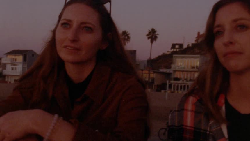 Two women are sitting on a beach near a coastal town at dusk