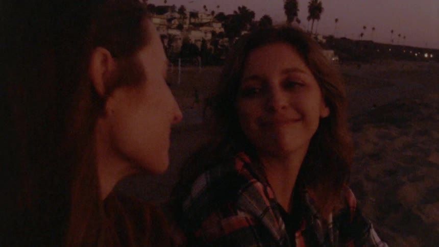 Two women are sitting on a beach by a coastal town and looking and smiling at each other