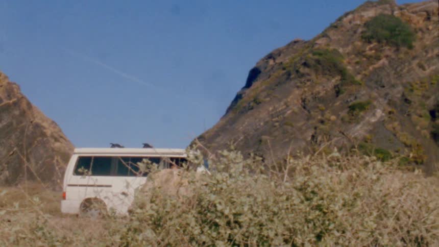 Two people park van near gap between two hills under a clear blue sky with a faint contrail