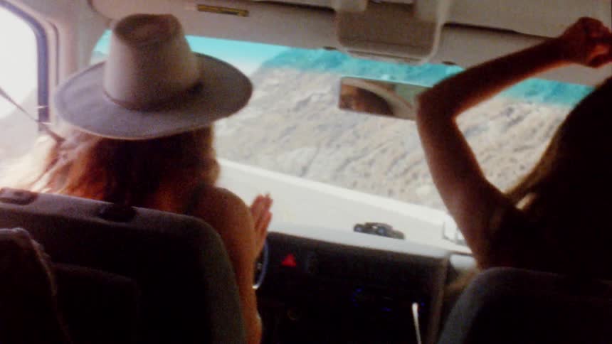 A woman driving along the shore taps her hands on the steering wheel as her friend sitting next to her dances