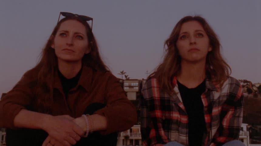 Two women sitting outside with buildings in background look forward before turning to share a smile