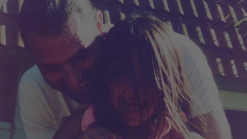 Father with long hair and white t shirt holds and kisses her daughter wearing pink dress in the garden