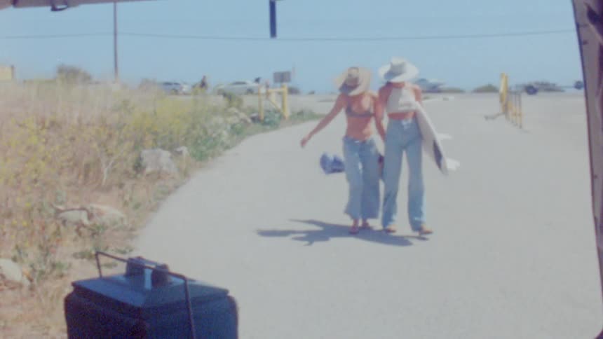 Two women walk back to their vehicle with a surfboard as one holds onto her hat