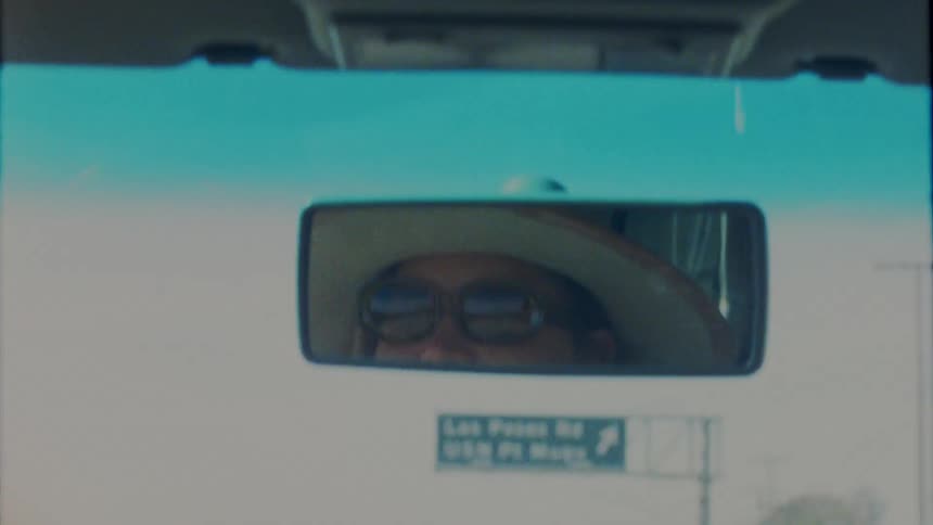 Face of a woman wearing a hat and sunglasses reflected on the rearview mirror of a moving car