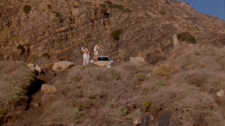 One woman stands on vehicle and another stands on ground nearby with mountain in the background