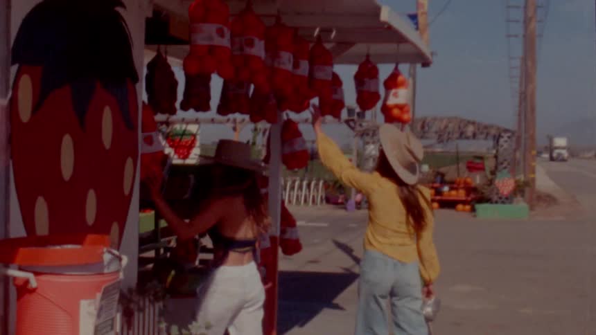 Two women walk towards a market on the side of the street and one woman reaches up to look at a bag of fruit