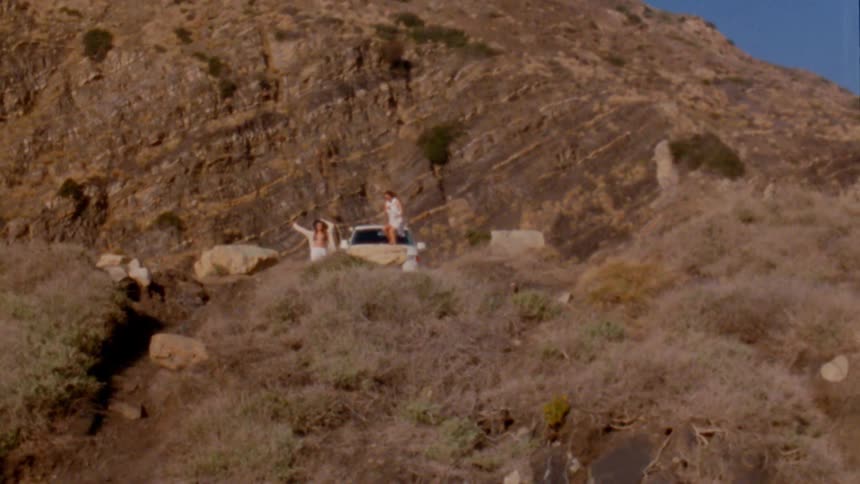 A car is parked on a mountain road and two people get out and a female jumps on the hood