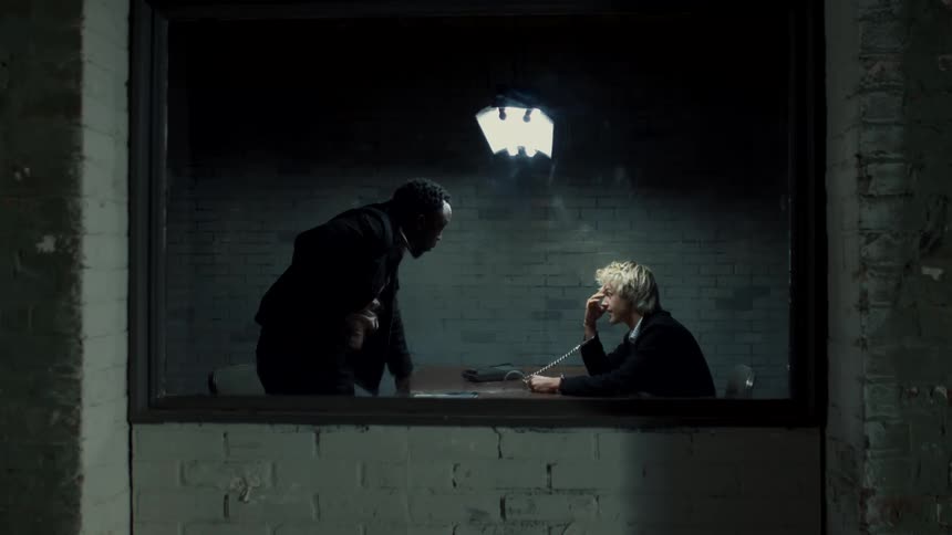A white man is handcuffed to a table and is being questioned by a black man