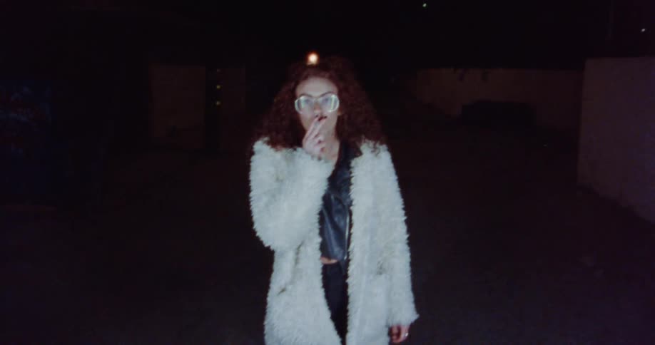 Female model with curly hair glasses black leather jacket and white feather jacket smokes cigarette outdoors at night