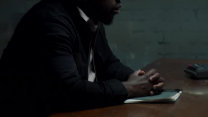 A man sitting with hands folded at a jail house table with a notebook and tape recorder