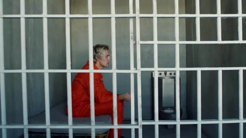 Blonde prisoner in prison uniform sits on bed in prison