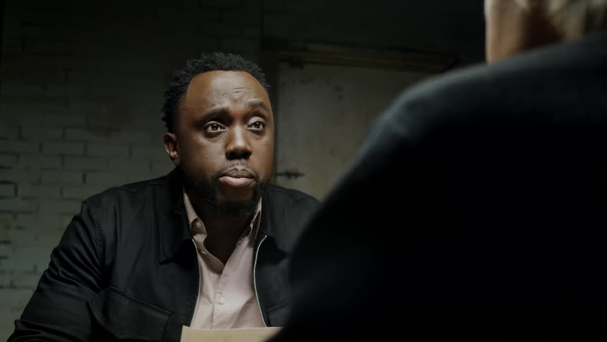 A black man is sitting inside a brick room talking a man