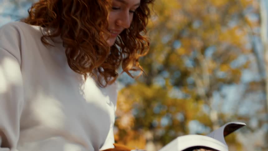 A young woman sitting beneath a tree grins while reading a book