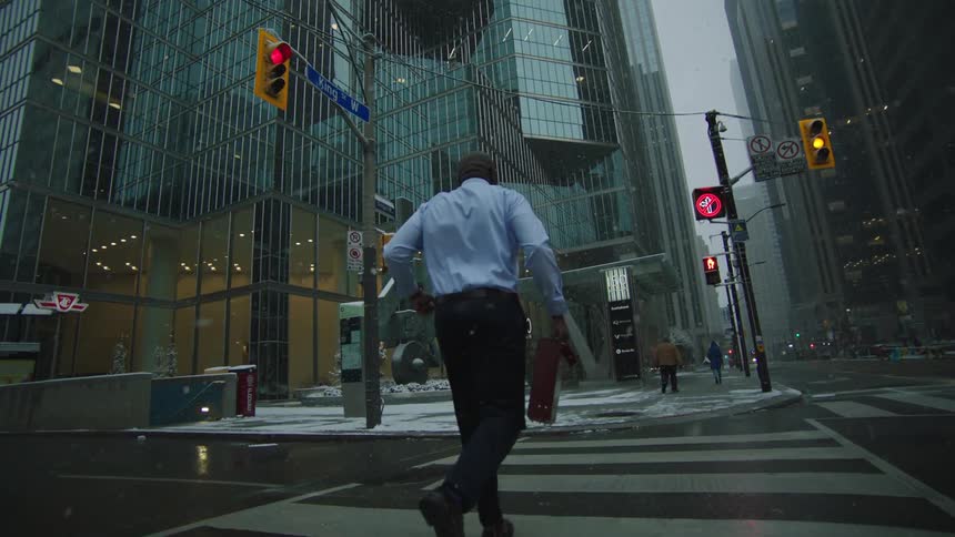 Businessman with blue shirt black pants and shoes carrying briefcase runs across the street with high rise office buildings as it rains