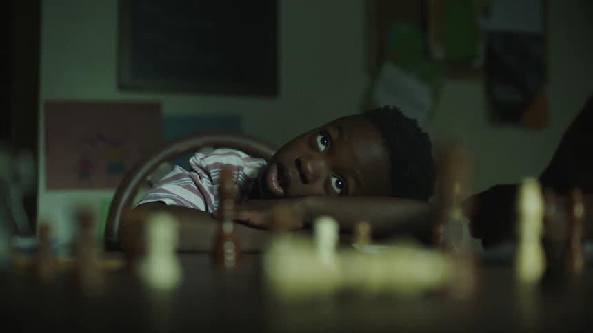 A finger knocks over the white king on a chess board while a boy looks on with head resting on his arms
