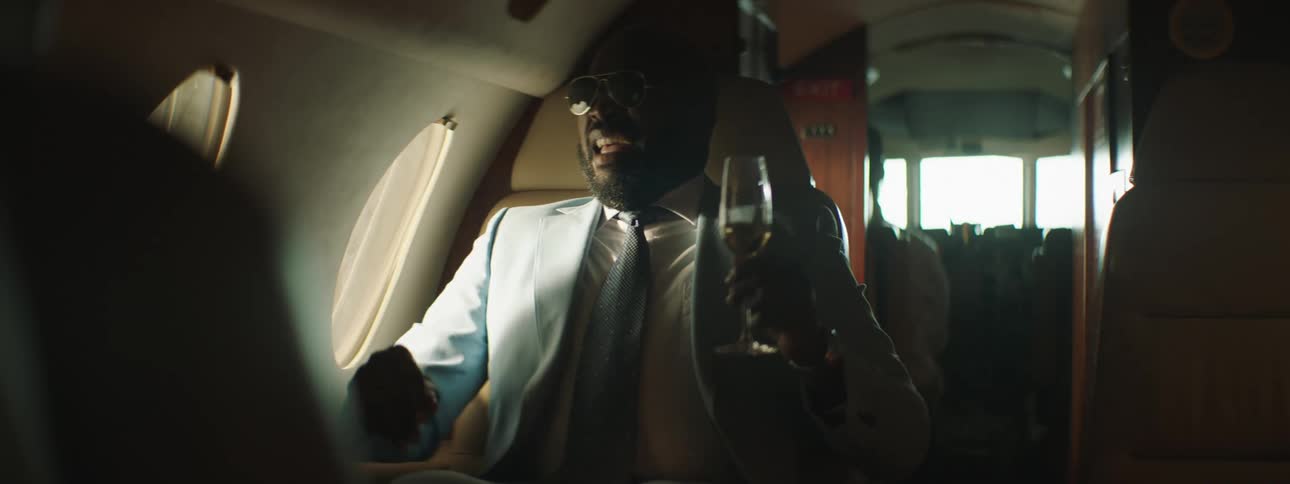 A man sitting on a private jet holding a champagne flute sneezes and spills his champagne everywhere