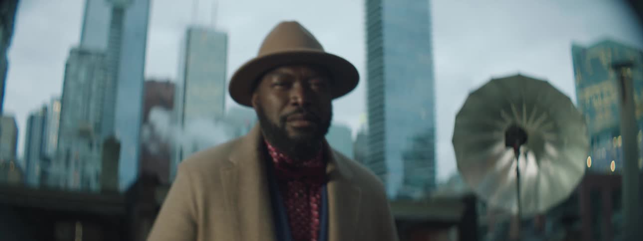 A black man is modeling a suit and hat and there are tall city buildings in the distance