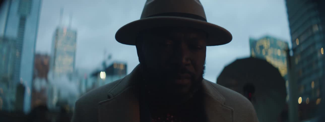 Man wearing a brown hat and coat surrounded by flashing lights in the city at evening