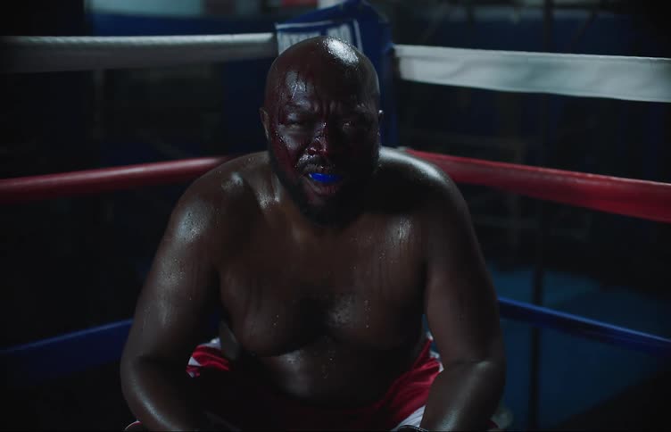 A boxer takes deep breaths while sitting on a stool in the ring as a camera flashes