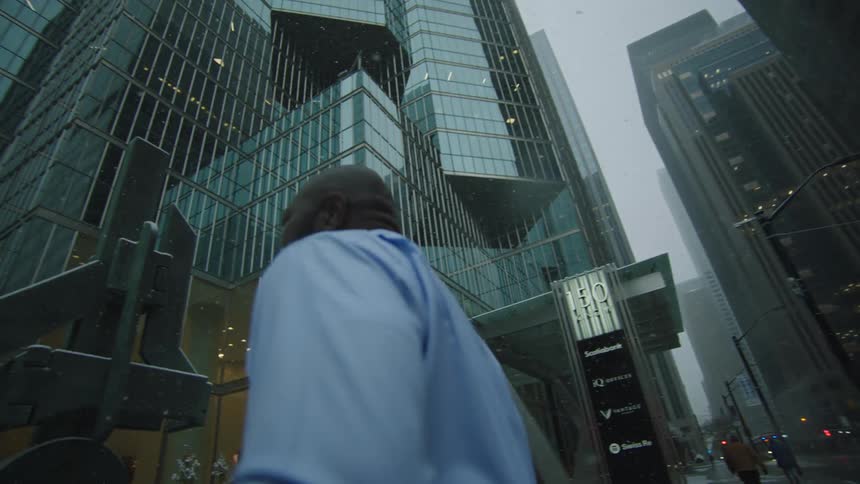 Businessman with beard blue shirt pants and tie carries briefcase looking around and running anxiously in the city street with high rise office buildings as it rains
