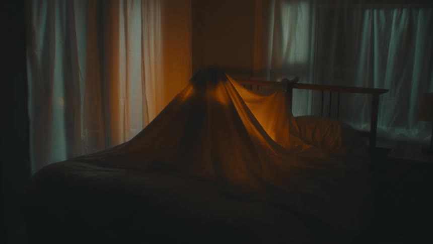 A silhouette of a person under a sheet that they have made in to a fort in a bed with a light shining inside