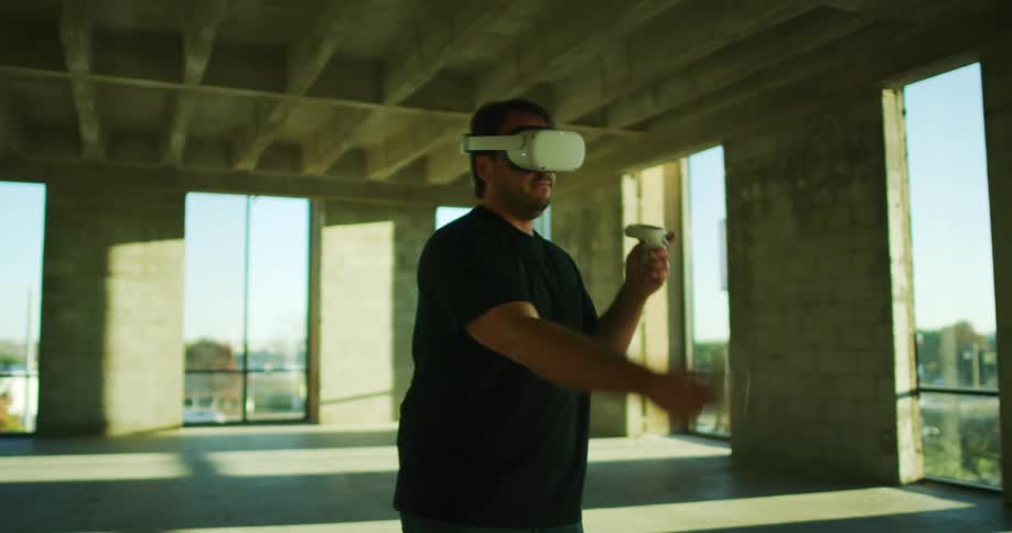 Man wearing virtual reality headset plays game using game controllers while standing in large empty room with busy highway visible through windows