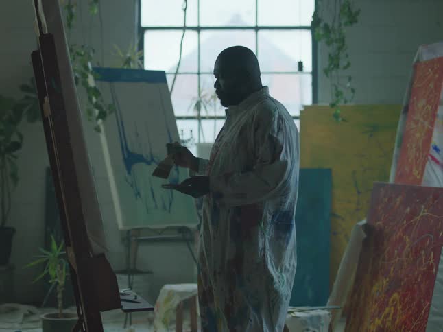 A black man is holding a paint palette and painting a picture