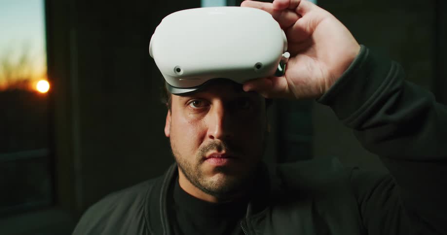 Man with brown hair eyes and black jacket takes off virtual reality headset and puts it back on in room at sunset