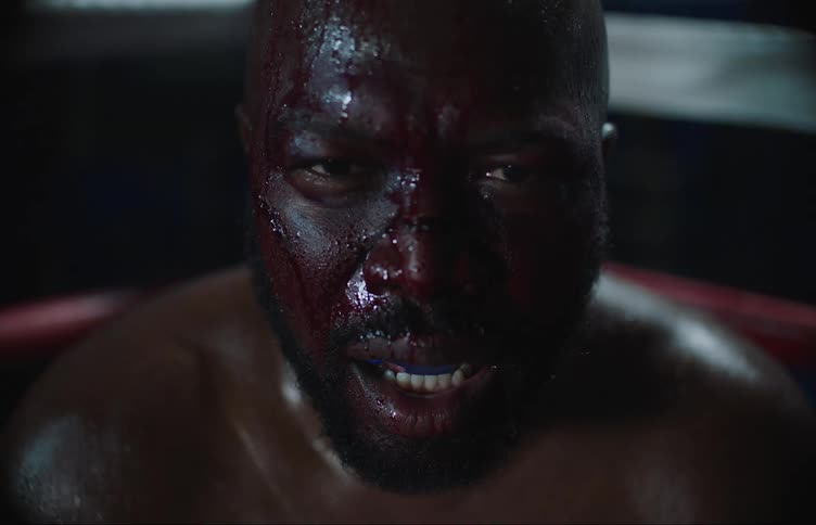 A black man with a bloody face is sitting in the corner of a boxing ring as camera flashes shine on his face