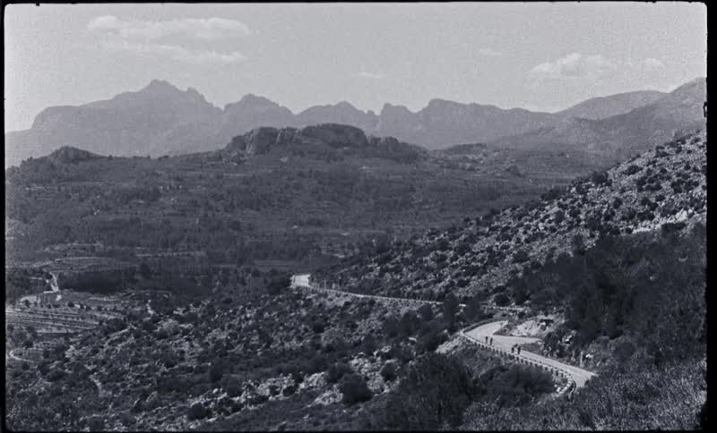 Black and white footage of cyclists driving on a road in the mountains caught on vintage film