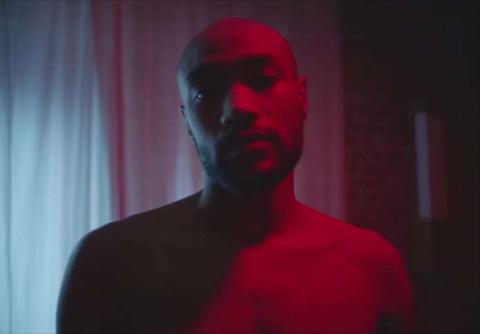 Bald and shirtless man with beard looks at camera in room with red lights