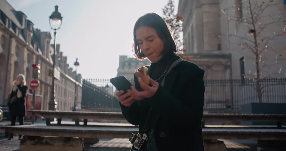 A young woman with a camera around her neck sits on a bench outside and eats as she looks at her cell phone