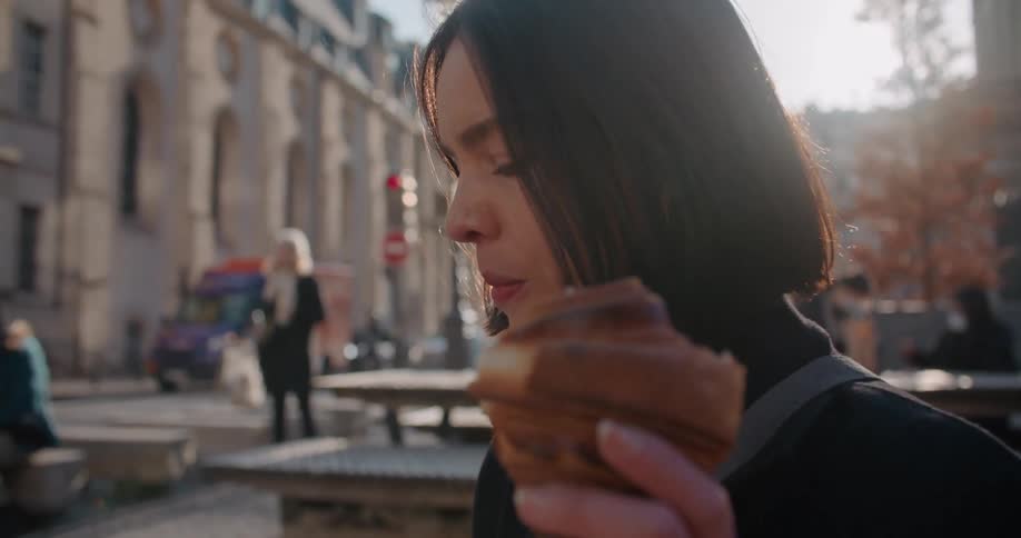 A woman eats a croissant while looking at her phone in a city