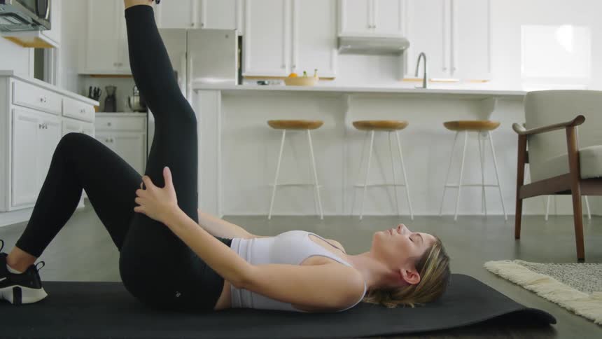 A woman lies on a mat in her home and stretches with her leg in the air