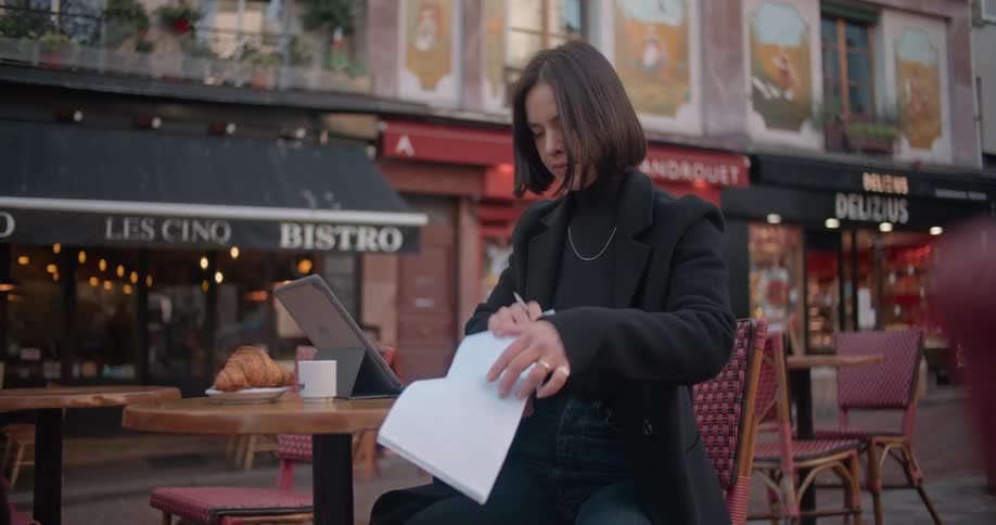 Woman with short straight hair black overcoat and sweater sits at cafeteria table turns over notebook pages next to tablet in the city street