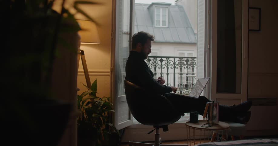 Man with beard and black turtleneck sweater working on laptop is silhouetted against the apartment balcony