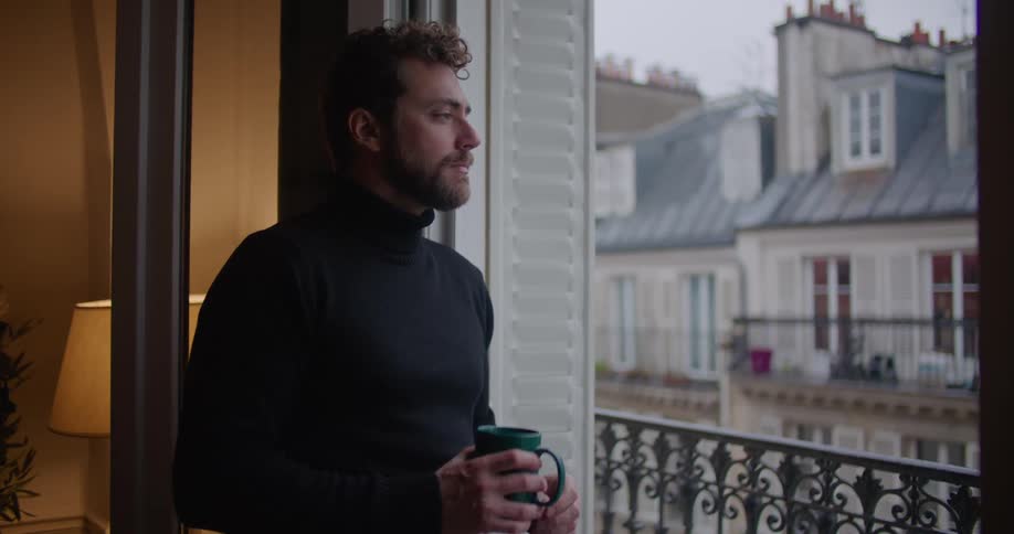 A man stands by a large open window drinking from a coffee mug
