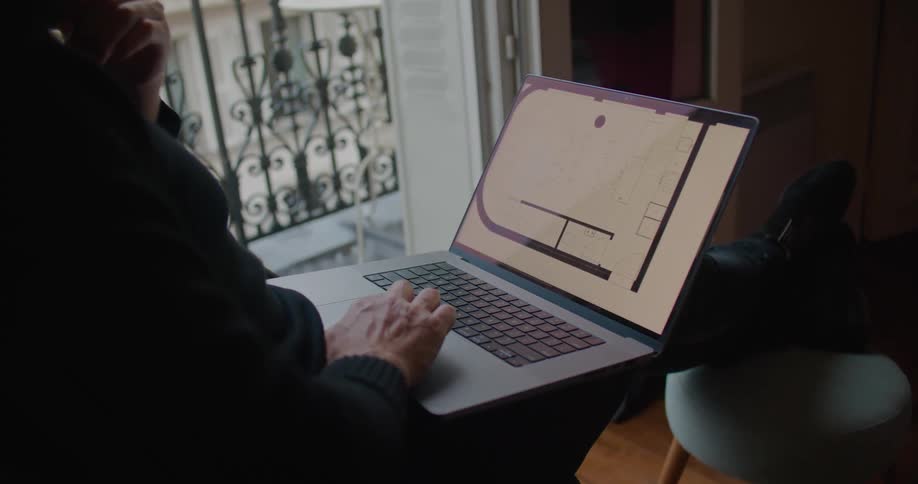 Young man wearing a black sweater working on a laptop near the apartment balcony