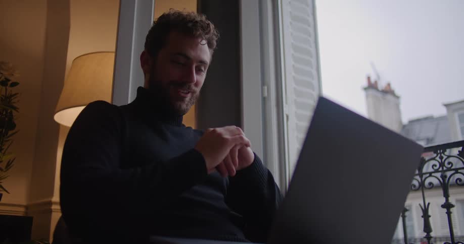Young man wearing a black sweater talking on a laptop near the apartment balcony