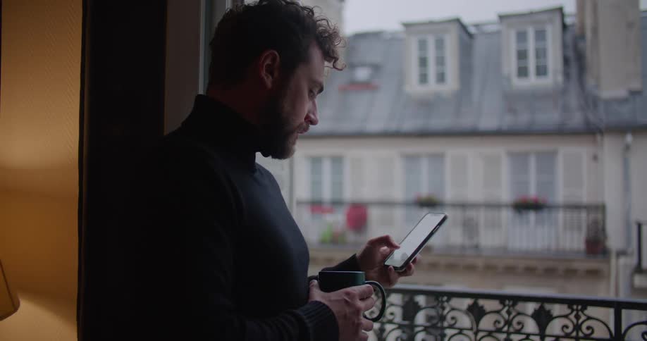 Young man wearing a black sweater holding a mug using his phone near the apartment balcony