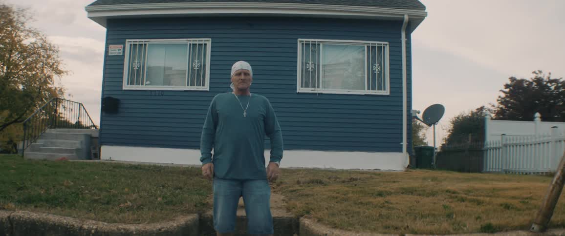 A man wearing a white bandana and shorts sits on the steps outside of a blue house and he stands up