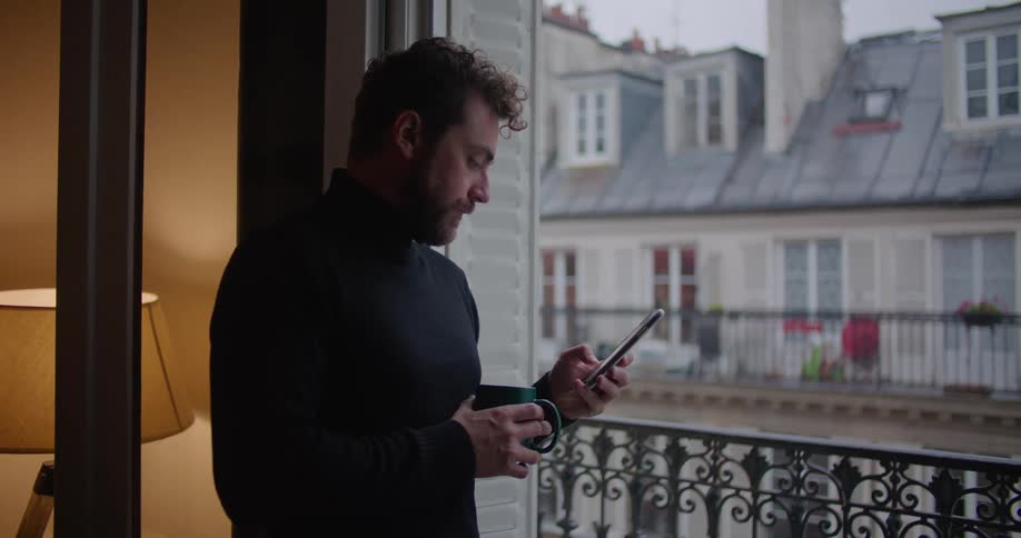 Young man wearing a black sweater holding a mug using his phone near the apartment balcony