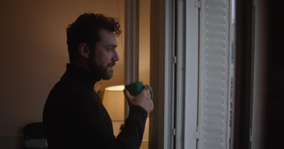 Man with beard and black turtleneck sweater drinks from green cup and looks out apartment balcony