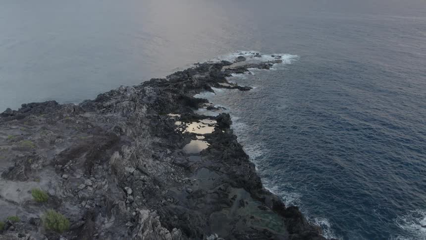 Aerial view of a rocky headland in the coast close to the ocean near mountains at evening