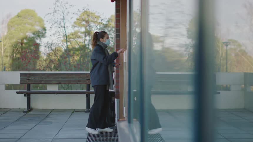 A woman wearing a face mask walks up to an outside glass door and swipes her key card and goes inside