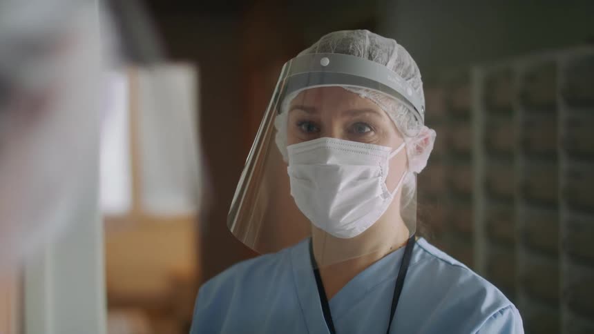 A nurse wearing a face mask and shield is nodding and talking to another person