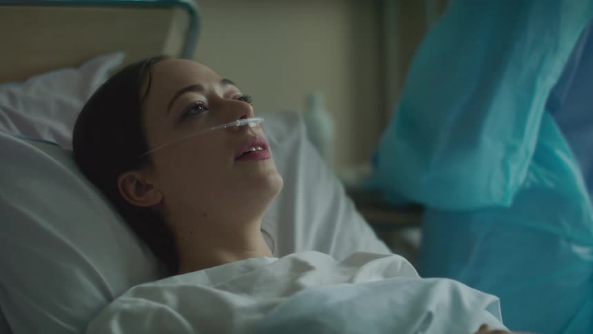 A woman with a breathing tube in her nose lies in a hospital bed with a nurse standing beside her